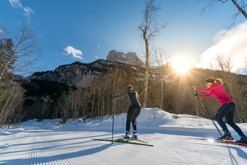 Two people cross-country skiing on a snowy trail, surrounded by mountains and trees.