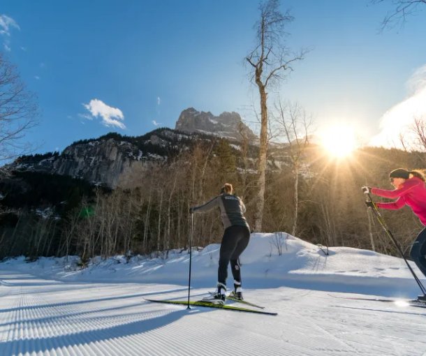 Zwei Personen beim Skilanglauf auf einer verschneiten Spur, umgeben von Bergen und Bäumen.