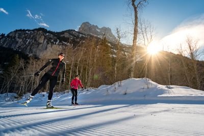 Two people cross-country skiing on a snowy trail, surrounded by mountains and trees.