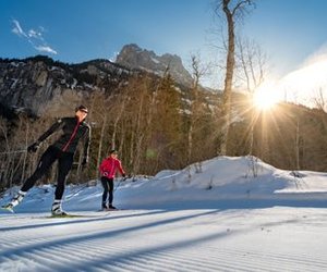 Zwei Personen beim Skilanglauf auf einer verschneiten Spur, umgeben von Bergen und Bäumen.