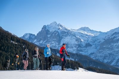 People with snowshoes and poles are hiking in a snowy mountain landscape.
