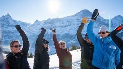 Personen mit Winterkleidung heben die Arme vor schneebedeckten Bergen in die Luft.