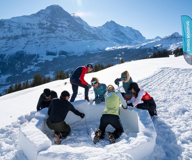 People are building an igloo in the snow, surrounded by mountains. Some are wearing winter clothing and hats.