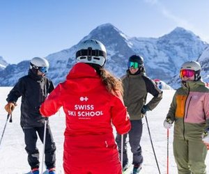 People with ski equipment and helmets are standing on a ski slope, surrounded by snow-covered mountains.