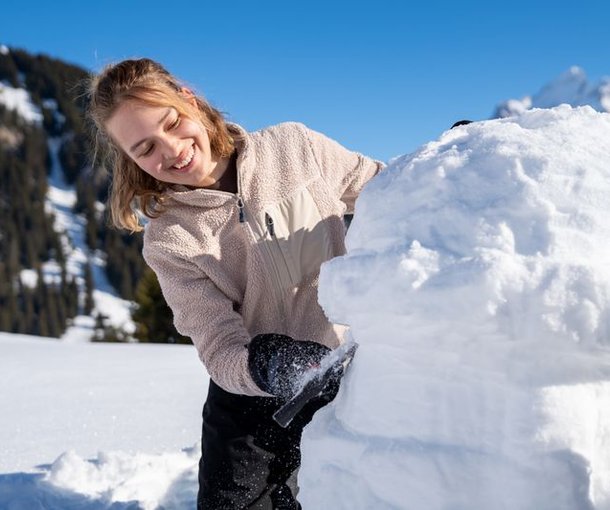 A person is building in the snow, wearing gloves; mountains in the background.