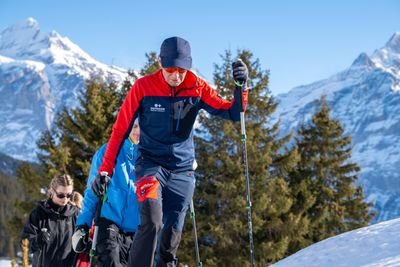 People with hiking poles and winter clothing walk in the snow in front of a mountain backdrop and trees.