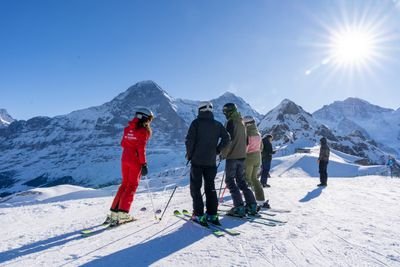 People with skis and helmets stand on a snow-covered mountain, surrounded by an Alpine landscape.
