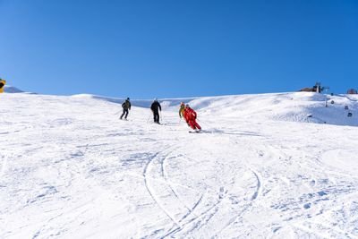 Drei Personen mit Skiern fahren einen schneebedeckten Hang hinunter, blauer Himmel im Hintergrund.