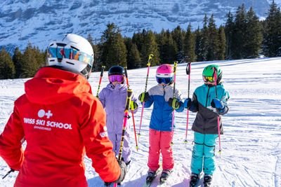Skilehrer in roter Jacke unterrichtet drei Kinder mit Helmen und Skiern auf verschneitem Berg.