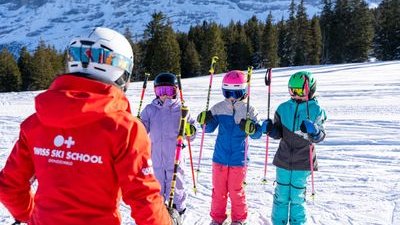 Skilehrer in roter Jacke unterrichtet drei Kinder mit Helmen und Skiern auf verschneitem Berg.