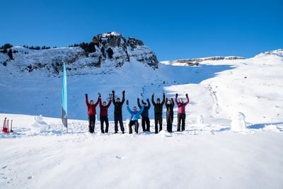 Group of people in winter clothing raising their arms in snow-covered mountain terrain in front of a rock face.
