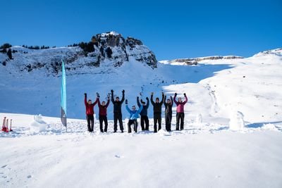 Group of people in the snow, raising arms, mountains in the background, blue flag on the left, winter clothing.
