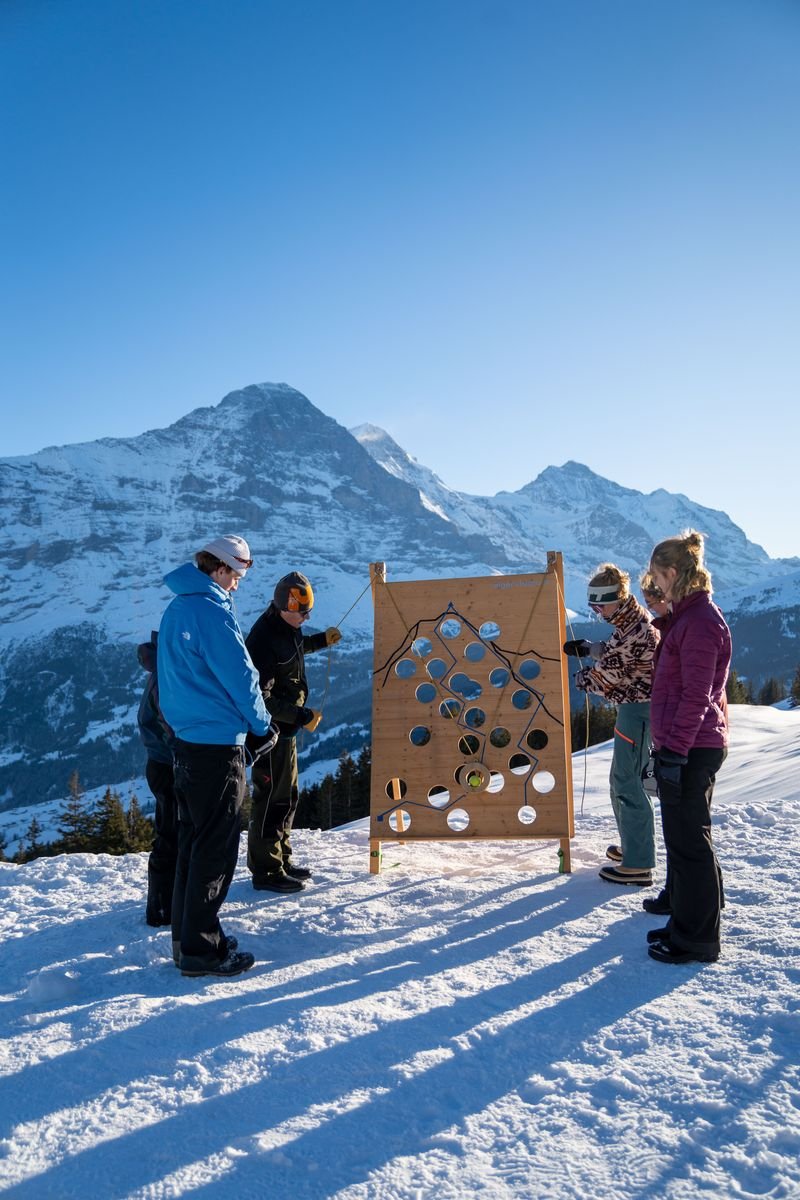 People are standing in the snow in front of a wooden board with holes, mountains in the background.