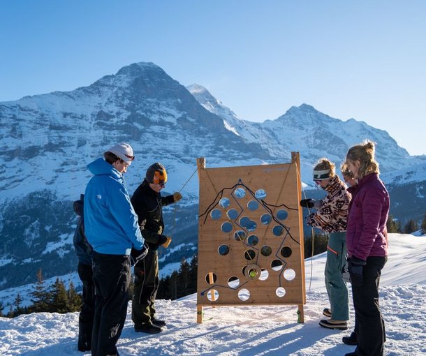 People are standing in the snow in front of a wooden board with holes, mountains in the background.