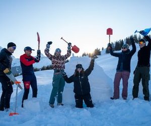 Six people with shovels in the snow, surrounded by mountains, wearing winter clothing and sunglasses.