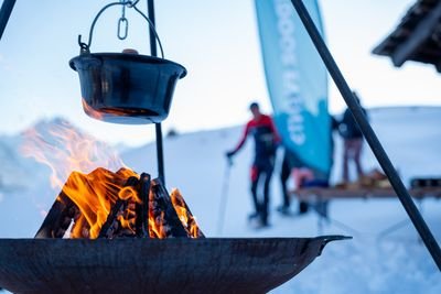 A cauldron hangs over a fire in the snow; in the background people with ski poles and a banner.