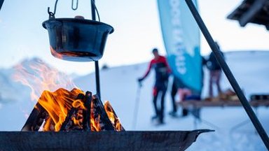 Ein Kessel hängt über einem Feuer im Schnee; im Hintergrund Personen mit Skistöcken und Banner.