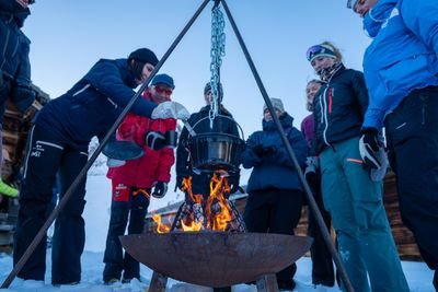 A group of people is standing in the snow around a campfire with a hanging kettle.