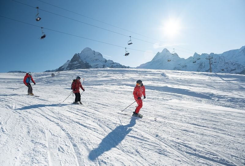 Skifahrer in Helmen und Ausrüstung fahren verschneite Hänge hinunter, im Hintergrund sind Berge und Skilifte zu sehen.