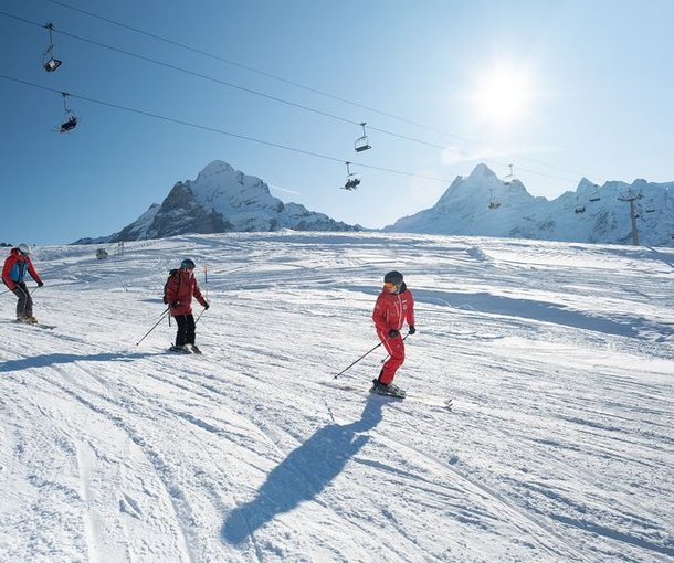Skiers in helmets and gear descend snowy slopes with mountains and ski lifts in the background.
