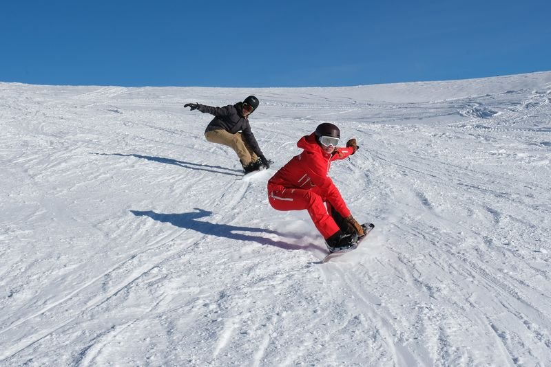 Zwei Snowboarder in Helmen und Schutzbrillen fahren unter einem klaren Himmel einen verschneiten Berghang hinunter.