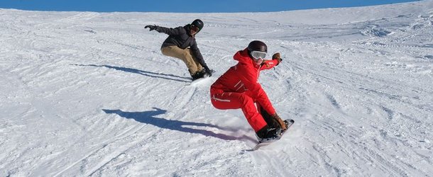 Two snowboarders in helmets and goggles ride down a snowy mountain slope under a clear sky.