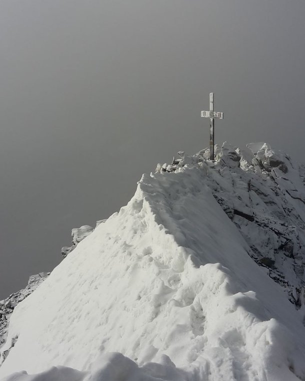 Snow-covered mountain peak with a cross, surrounded by foggy sky.