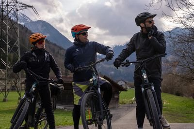 Drei Radfahrer mit Helmen und Sonnenbrillen auf Mountainbikes in einer ländlichen Gegend mit Blick auf Berge.