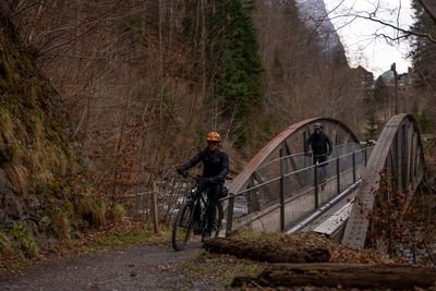 Cyclists wearing helmets ride across a bridge in a forested mountain area.