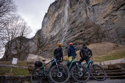 Three people with helmets and bicycles stand near a rocky cliff in a mountainous area.