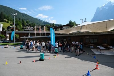 Gruppe von Menschen versammelt sich auf einem Platz mit Fahnen und Bergen im Hintergrund.