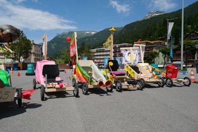 Bunte Seifenkisten stehen in einer Bergkulisse auf einem Parkplatz, umgeben von Verkehrskegeln.