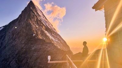 Person standing on a balcony at sunset, overlooking a mountain peak with snow patches.