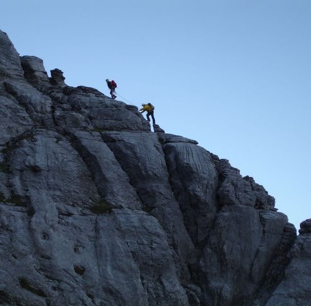 Two climbers in helmets and harnesses ascend a steep rocky mountain face, using ropes for safety.