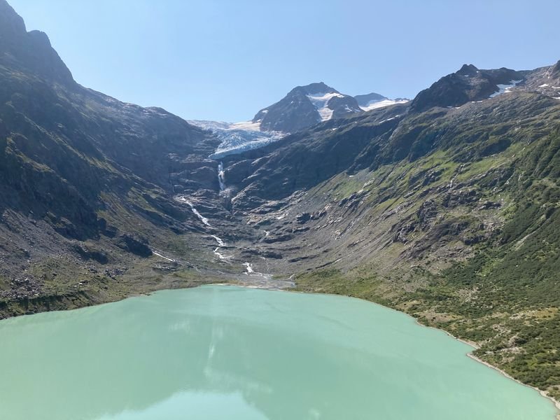View of a turquoise mountain lake, surrounded by green slopes and snow-capped peaks.