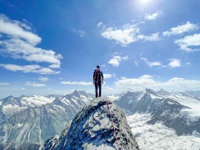 Person on mountain peak with climbing gear and rope, surrounded by snowy Alps.