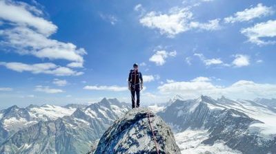 Person on mountain peak with climbing gear and rope, surrounded by snowy Alps.