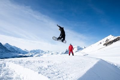 Snowboarder in der Luft bei einem Sprung auf schneebedecktem Hang, mit einer anderen Person in einem roten Anzug, der zuschaut, Berge im Hintergrund.