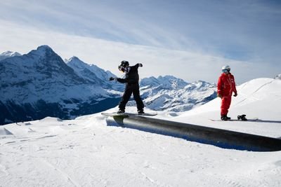Snowboarder in black gear on a rail, with a guide in red nearby, in a snowy mountain setting.