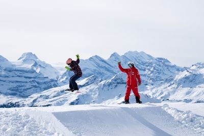Snowboarder jumping on snowy slope with instructor in red gear, set against mountain backdrop.