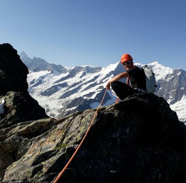 Climber in helmet and harness on rocky peak, snowy mountains in background, holding a rope.