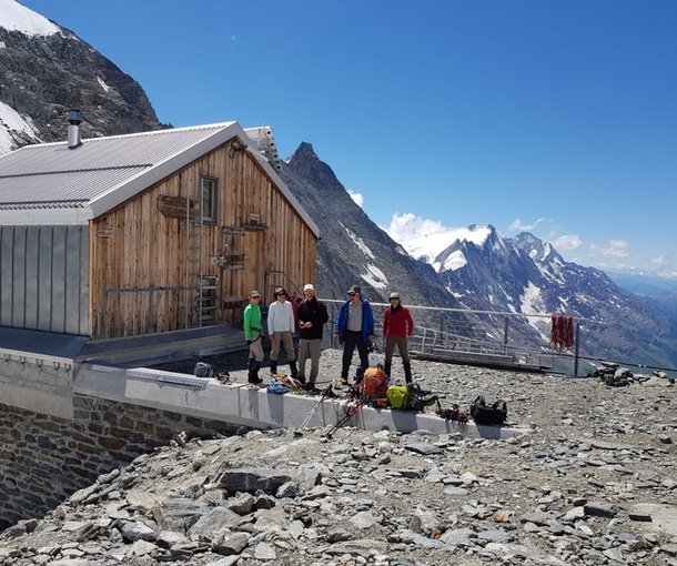 Four people with backpacks and climbing gear stand by a mountain hut, with snowy peaks in the background.