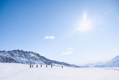 Group of people wearing winter clothing is hiking over snow-covered mountains under a clear sky.