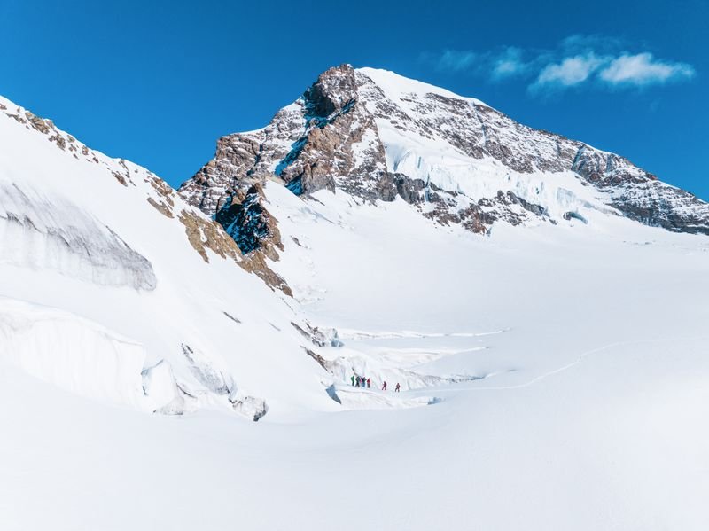 Personen mit Winterausrüstung wandern auf einem schneebedeckten Berggletscher unter blauem Himmel.