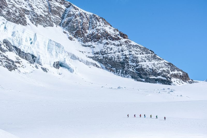 Personengruppe mit Winterkleidung wandert im Schnee vor einer verschneiten Bergkulisse.