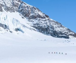Group of people wearing winter clothing hiking in the snow in front of a snow-covered mountain backdrop.
