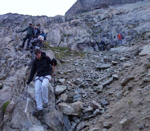 People with helmets and climbing harnesses are descending a rocky mountain path using poles.