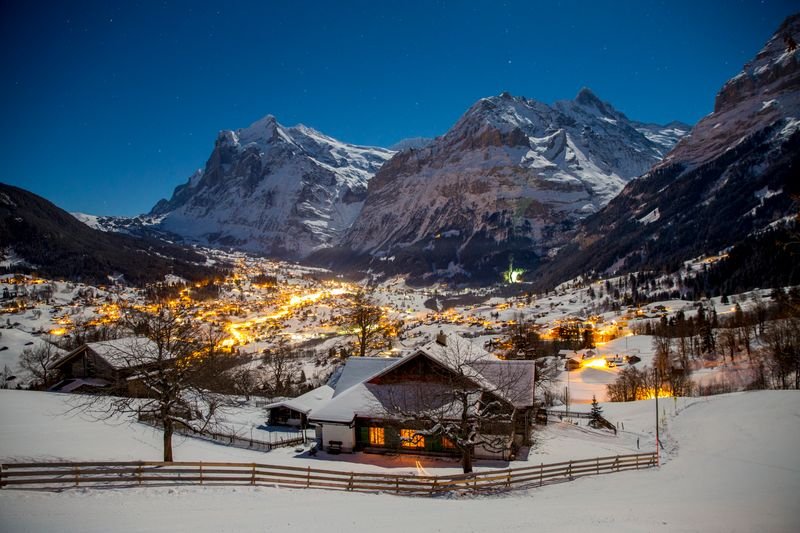 Snow-covered landscape with a lit-up village, surrounded by tall mountains at night.