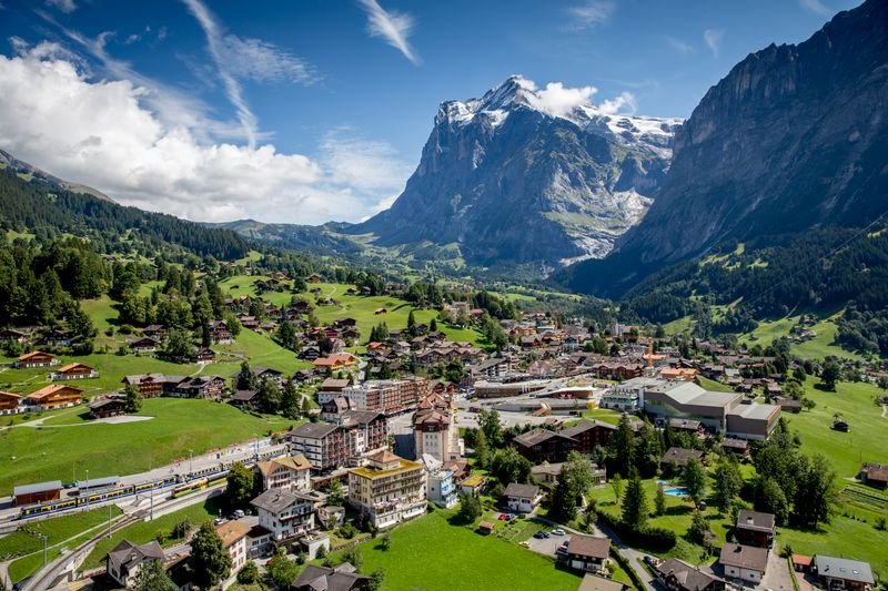 Aerial view of a village in the Swiss Alps with green meadows and snow-covered mountains.