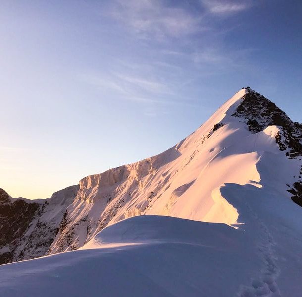Snow-covered mountain peak at sunrise with a clear sky, showing footprints along the ridge.